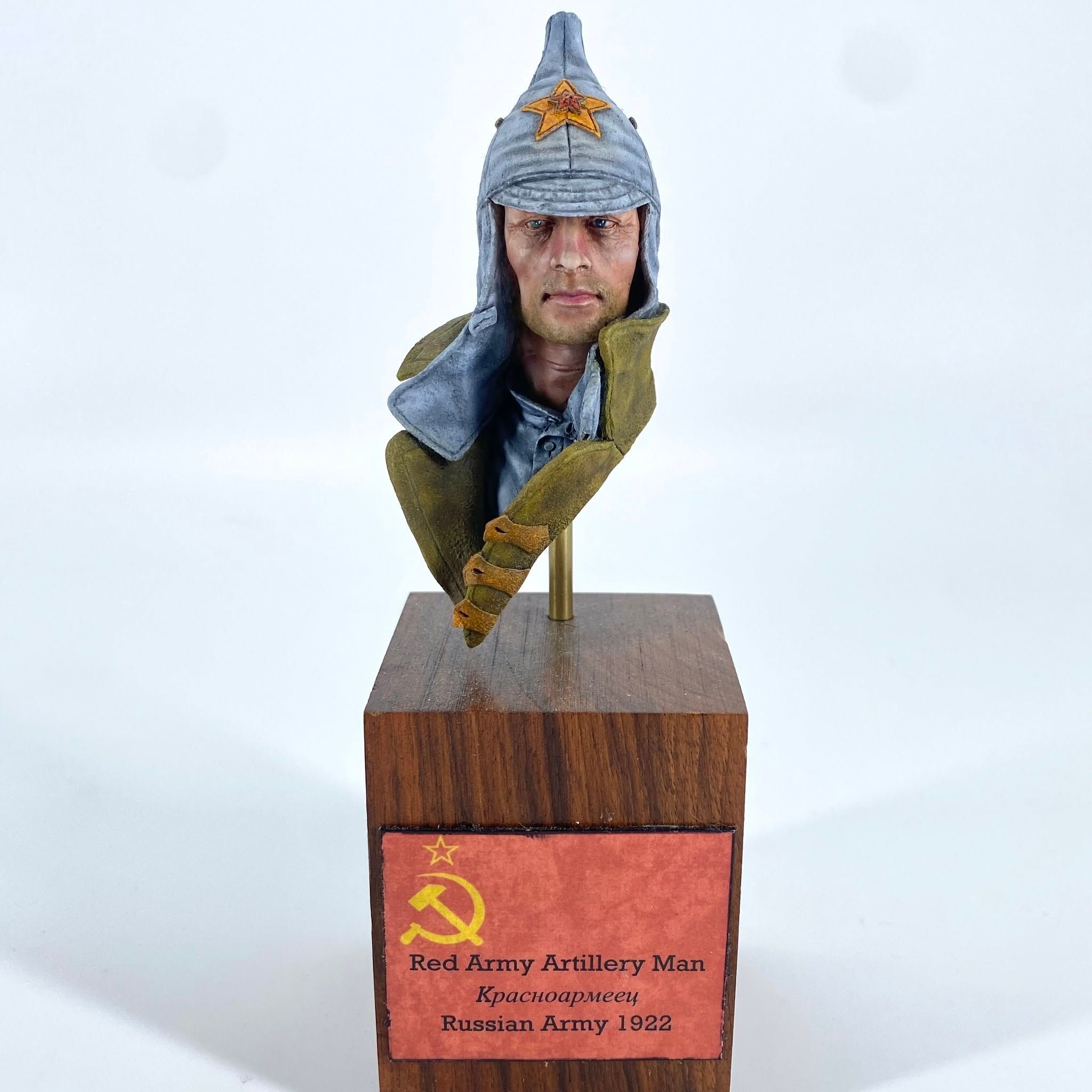 Painted bust of a Red Army soldier in a grey budenovka cap with a red star, wearing an olive greatcoat, mounted on a wooden plinth.