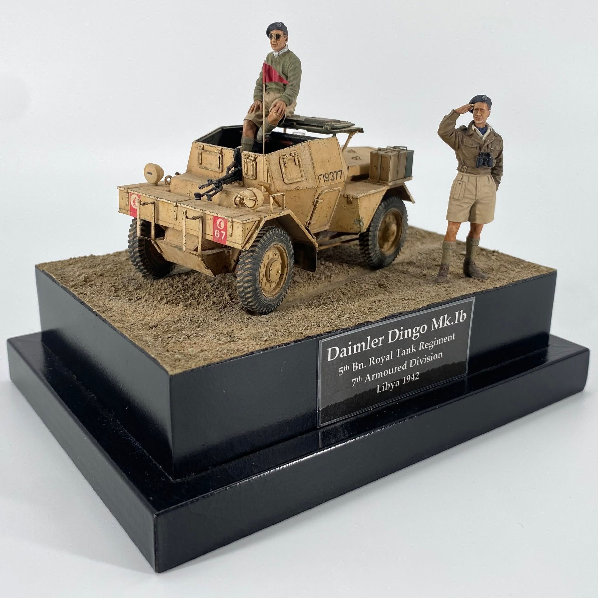Desert-sand scout car on a sandy base, a crewman seated on the hull and an officer in shorts saluting alongside.