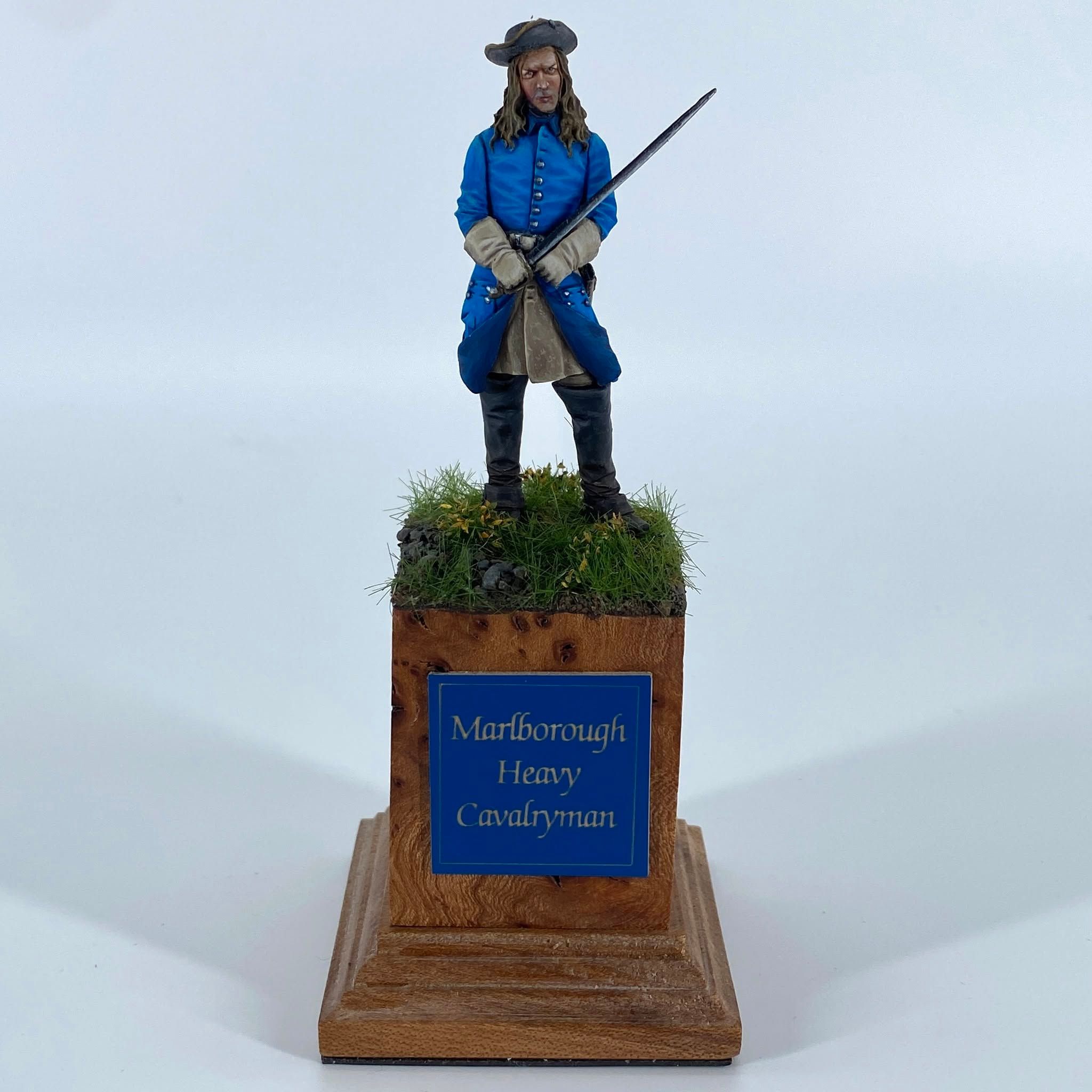 Standing figure of an early eighteenth century cavalryman in a bright blue coat and tricorne hat, holding a drawn sword, on a grassy base.