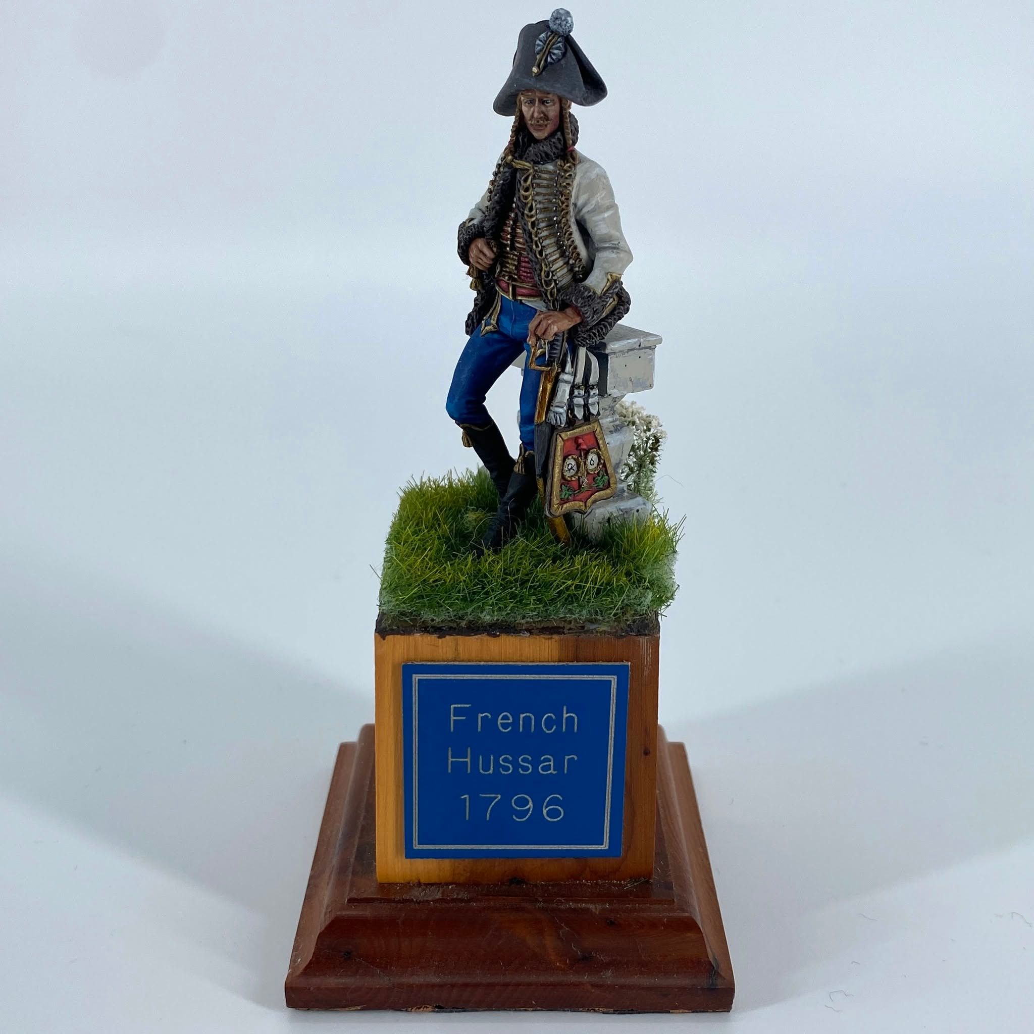 Figure of a French hussar in a bicorne hat, braided pelisse and blue breeches, leaning against a stone block on a grassy base.