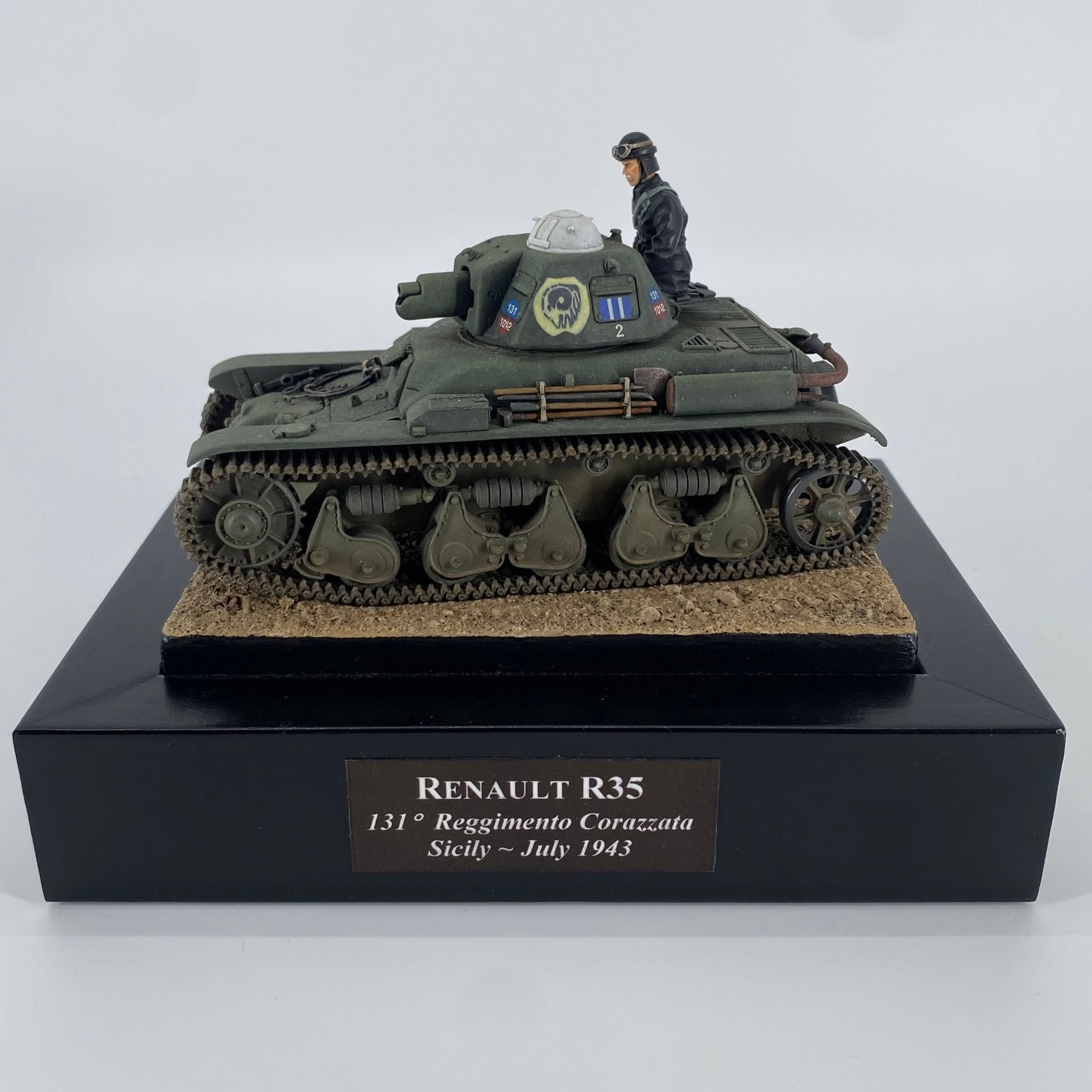 Olive green light tank with a rounded turret on a sandy base, a black-uniformed commander standing in the open hatch.
